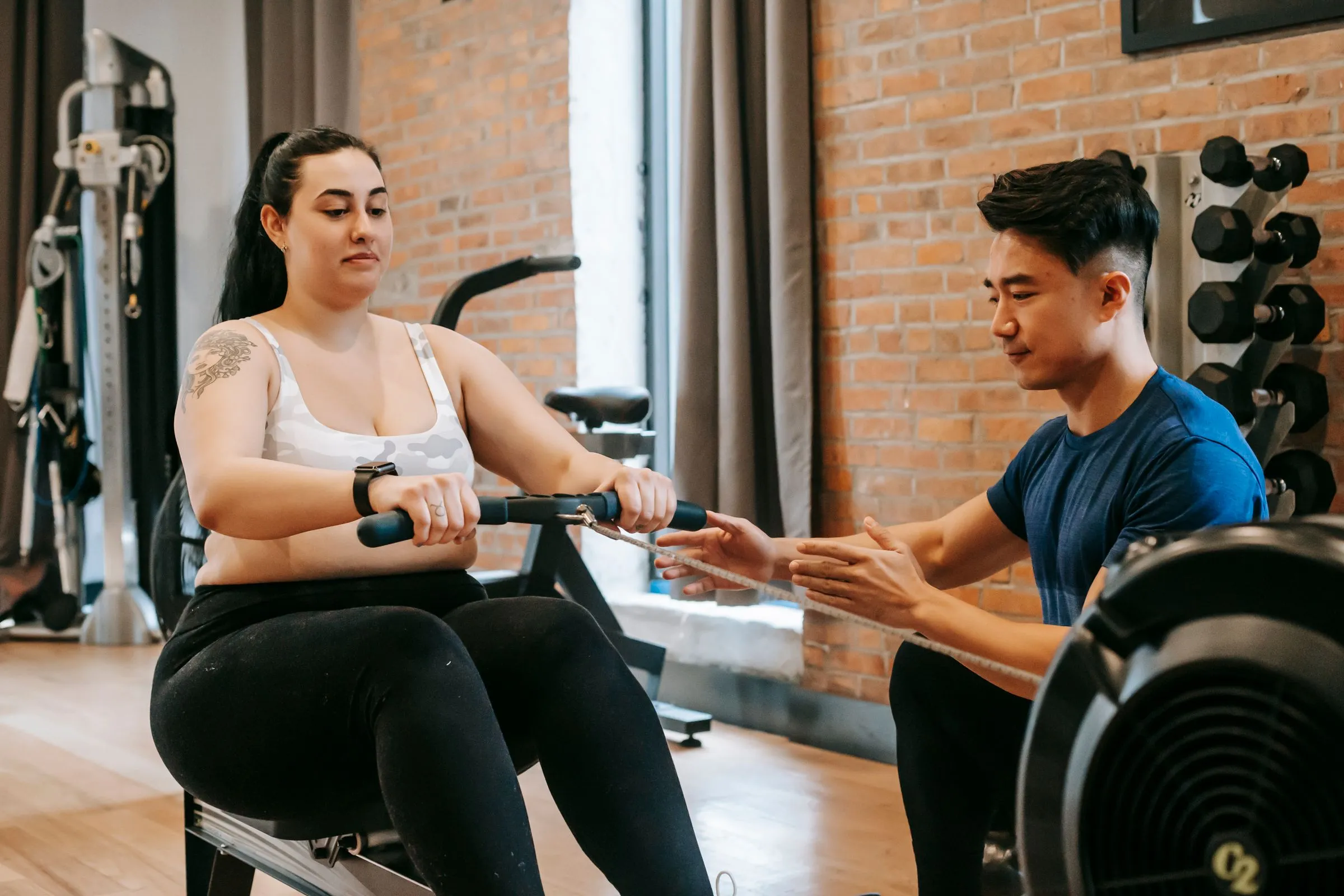 A coach crouching beside a member on a rowing machine, showing her how to hold the handle, in a brick-walled gym.
